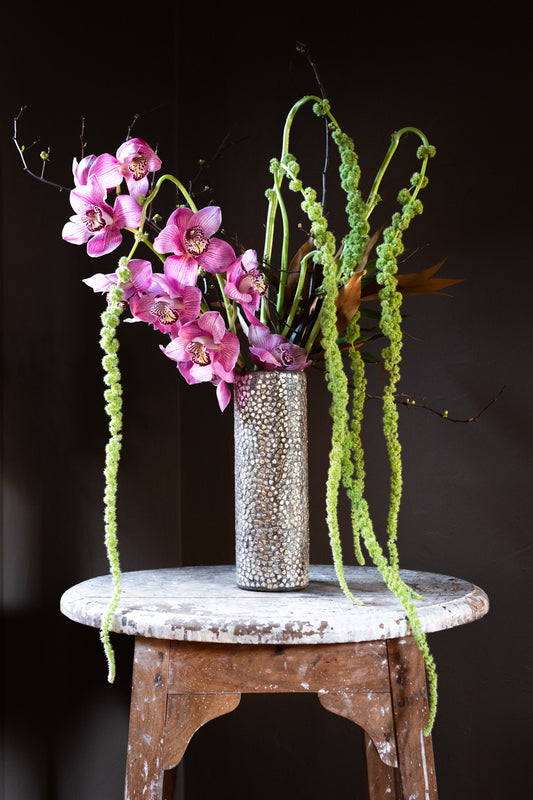 A modern and romantic flower arrangement featuring mauve cymbidium orchids, blooming quince branches, and hanging amaranthus designed in a tall silver hearts vase.  