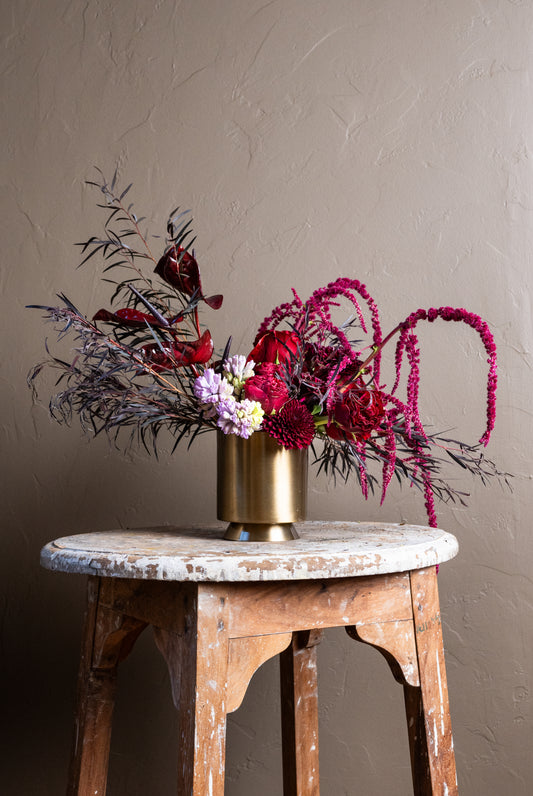 An organic and romantic flower arrangement featuring lavender hyacinth, red heart roses, pink amaranths, burgundy charm peonies, deep chrysanthemums and anthurium, all designed in a modern gold vase.