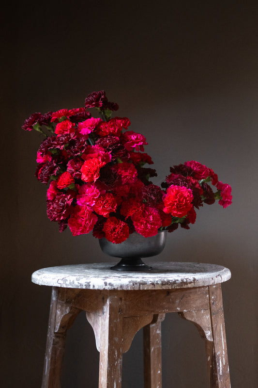 A dramatic and sexy flower arrangement featuring rich hues of deep burgundy, vibrant red, and bold hot pink carnations designed in a modern stacked style in a gunmetal compote vase